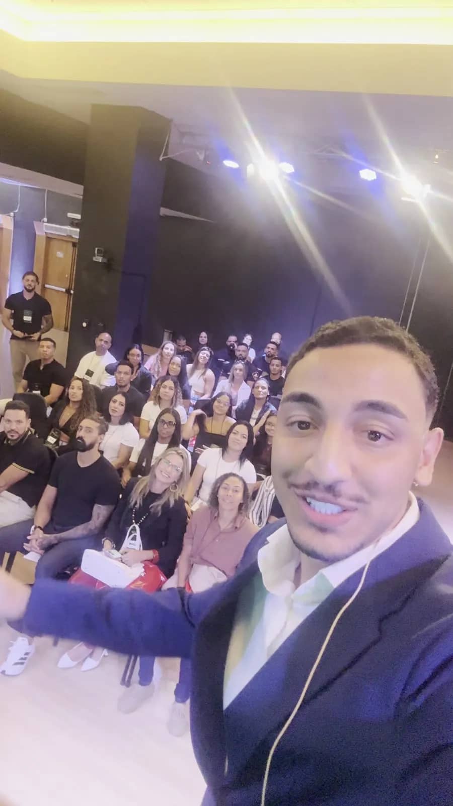 Thiago Máximo fazendo selfie no palco com o auditório cheio ao fundo, durante uma palestra